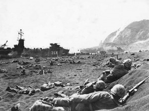 Marines burrow in the volcanic sand on the beach of Iwo Jima, as their comrades unload supplies and equipment from landing vessels despite the heavy rain of artillery fire from enemy positions on Mount Suribachi in the background. Image-U.S. Military