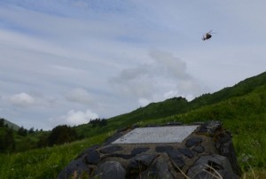 A Coast Guard MH-65 Dolphin helicopter flies over a plane crash memorial site on a hillside in the Anton Larsen Bay area to pay tribute during the 20th anniversary memorial service, June 26, 2015. The plane crash took the lives of three Coast Guardsmen and a civilian pilot, June 30, 1995. (U.S. Coast Guard photo by Petty Officer 3rd Class Lauren Steenson)
