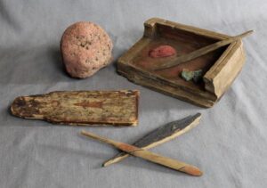 Photo: Objects associated with painting, Karluk One site, Koniag, Inc. Collection. Top row from left - Pumice grinder covered in red pigment, wooden bowl stained with red ochre, piece of red ochre, possible paint brush handle, piece of copper oxide; Bottom row from left - miniature skin sewing board painted with a sea otter, painted mask bangles (decorative attachments).
