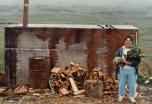 Photo: Rena Cohen outside an Akhiok steam bath, 1990. Photo by Priscilla Russell. KANA collection.
