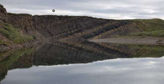 Part of the Nanushuk Formation, visible as a darker slanted rock layer, rises in a bluff above a bend of the Kukpowruk River. The formation, which is 800 to 5,000 feet thick across the central and western North Slope of Alaska, dates to the Albian-Cenomanian Age, roughly 94 million to 113 million years ago. A large number of dinosaur tracks were found in this area.
